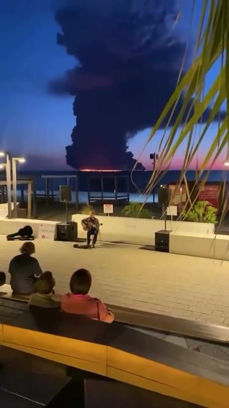 Street Musician Performs Against Backdrop of Burning Tanker